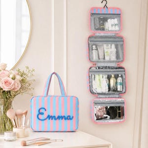 May include: A hanging toiletry bag with four clear zippered compartments and a matching rectangular travel case, both featuring pink and blue vertical stripes. The travel case displays the name Emma in blue script lettering on the front.