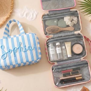 May include: A light blue and white striped toiletry bag with the name Sophia in raised blue lettering. Beside it, an open pink and white striped hanging toiletry bag features multiple clear zippered compartments filled with various cosmetics.