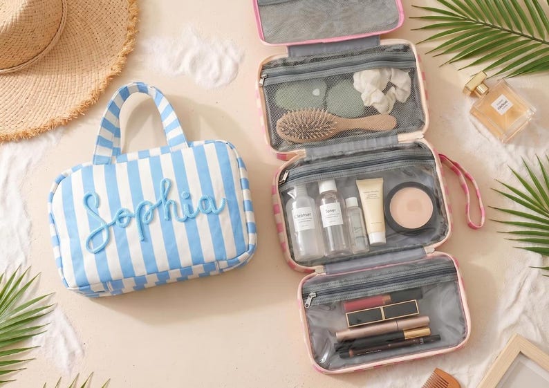 May include: A light blue and white striped toiletry bag with the name Sophia in raised blue lettering. Beside it, an open pink and white striped hanging toiletry bag features multiple clear zippered compartments filled with various cosmetics.