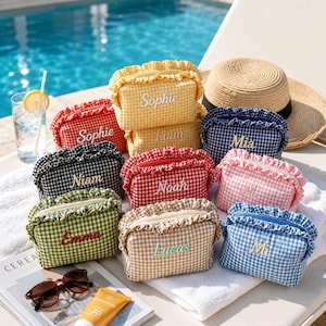 May include: A collection of small, rectangular zippered pouches featuring a gingham pattern in various colors including yellow, red, black, green, beige, pink, and blue. Each pouch has a ruffled trim and is embroidered with a unique name.