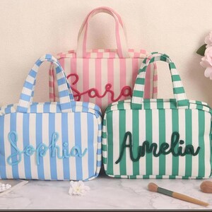 May include: Three rectangular zippered pouches with handles, each featuring vertical stripes and a name in cursive script. One pouch is pink and white with the name Sara, one is blue and white with Sophia, and one is green and white with Amelia.