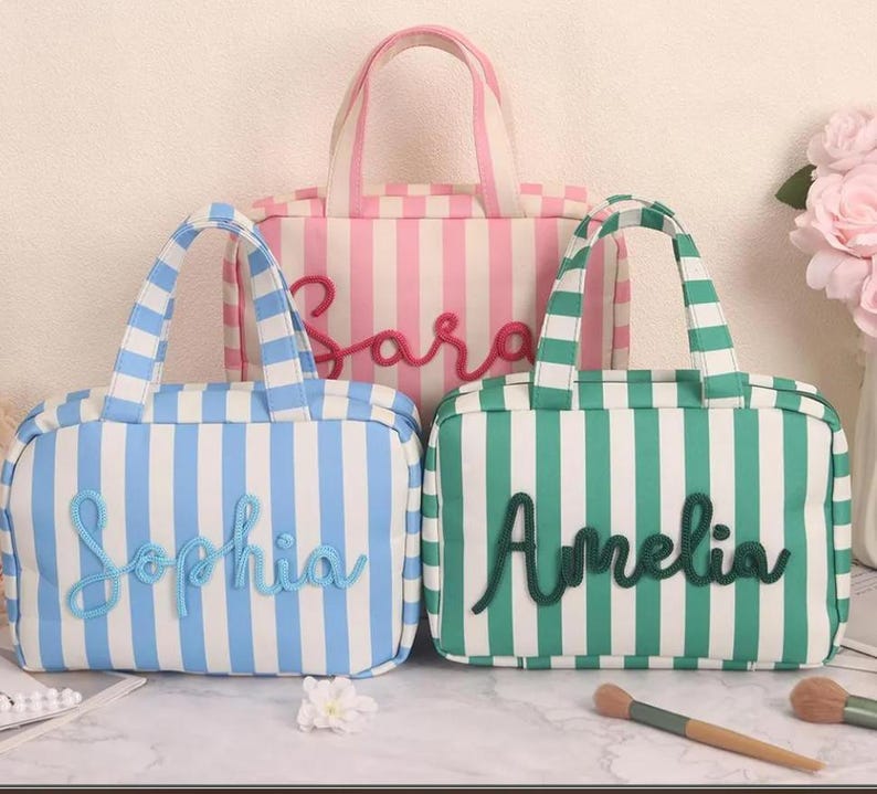 May include: Three rectangular zippered pouches with handles, each featuring vertical stripes and a name in cursive script. One pouch is pink and white with the name Sara, one is blue and white with Sophia, and one is green and white with Amelia.