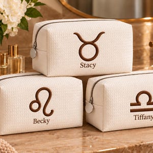 May include: Three rectangular, off-white textured cosmetic bags with silver zippers. Each bag features a brown embroidered zodiac symbol and a name: Taurus with Stacy, Leo with Becky, and Libra with Tiffany. The bags have a structured, boxy shape.