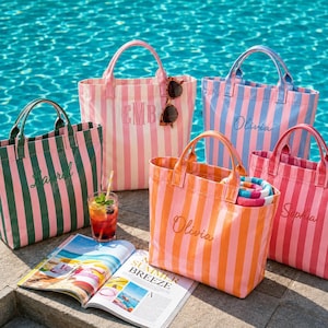Könnte beinhalten: Fünf gestreifte Tragetaschen in verschiedenen Farben wie Grün, Rosa, Blau und Orange, jeweils mit gestickten Namen oder Initialen. Die Taschen haben zwei Griffe und stehen neben einer offenen Zeitschrift und einem Glas mit einem Eiskaltgetränk.