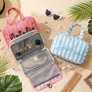 May include: Two rectangular striped travel bags with top handles and embroidered names. One bag is orange and pink with the name Natalia, and the other is blue and white with the name Sophia. The bags feature interior mesh pockets and elastic loops.