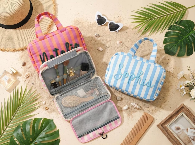 May include: Two rectangular striped travel bags with top handles and embroidered names. One bag is orange and pink with the name Natalia, and the other is blue and white with the name Sophia. The bags feature interior mesh pockets and elastic loops.