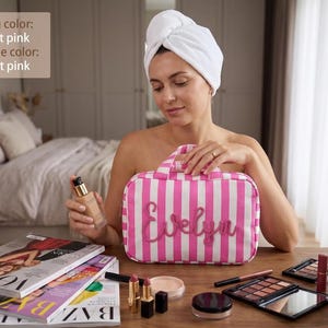 May include: A rectangular hot pink and white striped toiletry bag featuring the name Evelyn in hot pink script lettering on the front, with a top handle and a zipper closure, displayed alongside various makeup products and magazines.