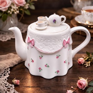 May include: A white ceramic teapot featuring a scalloped lid adorned with a miniature teapot and two tiny teacups. The body is decorated with small pink rose patterns, a textured lace-like upper border, and three-dimensional pink bows.