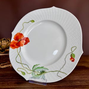 May include: A white porcelain plate with a scalloped edge and a textured wavy pattern on the rim. The plate features a hand-painted design of vibrant red poppy flowers, green stems, and leaves extending across the surface of the white ceramic.