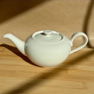 Könnte beinhalten: Eine weiße Teekanne aus Keramik mit rundem Körper, gebogenem Ausguss und passendem Deckel mit kleinem Knaufgriff. Die Teekanne hat eine glatte, glänzende Oberfläche und ein schlichtes, funktionales Design mit einem stabilen Griff.