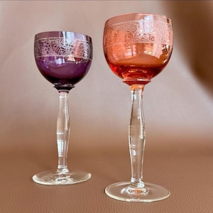 Könnte beinhalten: Zwei Stielgläser mit klaren Glasfüßen und Stielen. Ein Glas hat eine tiefviolette Kuppa, während das andere eine leuchtend orangefarbene Kuppa aufweist. Beide Kuppen sind mit einem geätzten Band mit detailliertem Weinrebenmuster verziert.