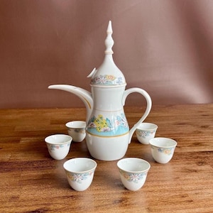 Könnte beinhalten: Ein Kaffeeservice aus weißem Porzellan mit einer hohen, schlanken Kanne im Dallah-Stil mit spitzem Deckel und fünf passenden kleinen Tassen. Das Set ist mit einem zarten Blumenmuster in Blau-, Gelb- und Rosatönen sowie Goldrändern verziert.