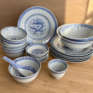 May include: A collection of white porcelain dinnerware featuring blue dragon motifs and translucent rice grain patterns. The set includes various bowls, plates, small dishes, a spoon, and a cup, all decorated with blue geometric border designs.