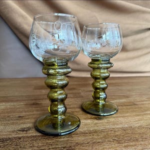 Può includere: Due calici in vetro trasparente con motivi floreali incisi sulla coppa. Ogni calice presenta uno stelo spesso in vetro verde oliva con una serie di segmenti arrotondati e bulbosi che terminano in un'ampia base circolare dello stesso vetro.