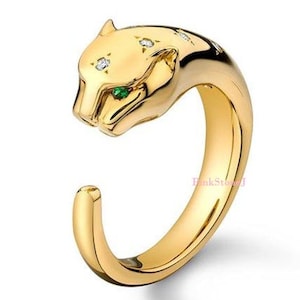 May include: A gold-toned open ring shaped like a panther head. The panther features a bright green eye and several small, clear, sparkling stones embedded along the top of its head. The band is smooth, polished, and curves into an open design.
