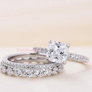 May include: A silver-toned engagement ring set featuring a cushion-cut centre stone with a pavé band, paired with a matching eternity band adorned with a row of round-cut stones. The text PinkStone is visible on the band of the engagement ring.