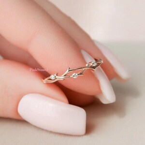 Peut inclure: Une bague en or rose présentant un design de bande délicat en forme de branche avec de petites épines texturées. La bande est ornée de trois petites pierres rondes et transparentes serties à intervalles le long de la structure métallique.