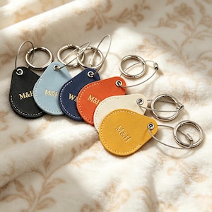 May include: A collection of six teardrop-shaped keychains in black, light blue, navy, orange, cream, and yellow. Each keychain features a textured surface, white stitching along the edges, and the letters M&H embossed in gold, attached to a metal ring.