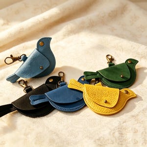 Könnte beinhalten: Eine Sammlung von vier vogelgeformten Schlüsselanhängern in Blau, Schwarz, Dunkelblau und Gelb. Jeder Anhänger verfügt über eine strukturierte Oberfläche, einen Metalldruckknopfverschluss und einen bronzefarbenen Karabinerhaken zur Befestigung.
