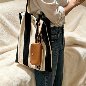 May include: A black and white vertical striped canvas tote bag featuring a small tan leather-like pouch attached by a thin cord. The pouch has a gold-toned snap button closure and is positioned over a light-colored smartphone case.