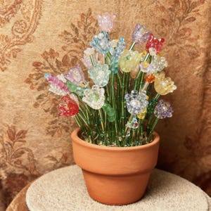 Puede incluir: Un arreglo decorativo de flores hechas con cuentas multicolores en una maceta de color terracota. Las flores presentan cuentas translúcidas en tonos rojo, azul, amarillo, morado y transparente, unidas a tallos de alambre verde.