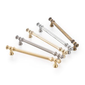 May include: A set of five metal cabinet pulls arranged in a diagonal row. The pulls feature a cylindrical bar design with rounded ends and two mounting posts. The finishes include brushed gold, matte silver, polished gold, chrome, and antique brass.