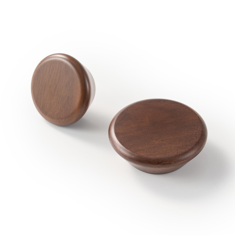 Op de afbeelding: Twee ronde kastknoppen gemaakt van donkerbruin hout. Elke knop heeft een glad, afgerond bovenoppervlak met een taps toelopende basis, met een natuurlijke houtnerftextuur en een gepolijste afwerking, gezien vanuit een hoge hoek.