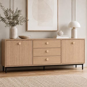 Op de afbeelding: Een dressoir van licht eikenhout met twee zijkasten met verticaal gegroefde deurpanelen en drie centrale lades. Elke deur en lade is voorzien van een kleine, ronde, bijpassende houten knop. Het meubelstuk staat op dunne zwarte metalen poten.