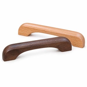 Puede incluir: Dos tiradores de armario arqueados hechos de madera maciza. Un tirador presenta una veta de madera natural de color tostado claro, mientras que el otro es de una madera marrón más oscura y rica. Ambos tienen un diseño curvo y suave.