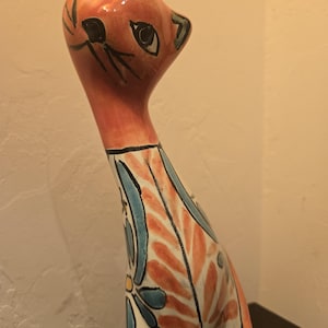 May include: A ceramic cat figurine with a long, slender neck and head turned to the side. The cat features a glossy orange finish on the head and neck, while the body is decorated with hand-painted blue, orange, and white floral patterns.