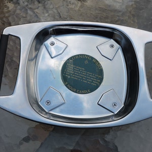 May include: A silver metal candle warmer with black handles. The warmer has four triangular indentations and a sticker that reads "Corning Ware 8 Hour Candle" with instructions for best results.