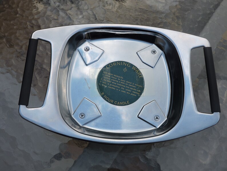 May include: A silver metal candle warmer with black handles. The warmer has four triangular indentations and a sticker that reads "Corning Ware 8 Hour Candle" with instructions for best results.