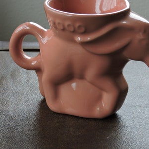 May include: A pink ceramic mug shaped like a donkey. The mug has a handle and a wide opening. The word "COOO" is printed on the side of the mug.