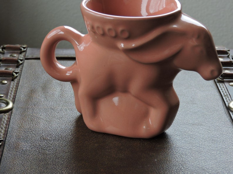 May include: A pink ceramic mug shaped like a donkey. The mug has a handle and a wide opening. The word "COOO" is printed on the side of the mug.