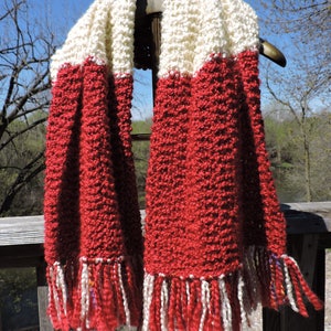May include: A handmade, textured scarf in red and cream. The scarf features a thick, knitted design with a fringed edge. The top section is cream, transitioning to red. The scarf is displayed outdoors, hanging on a decorative hanger.