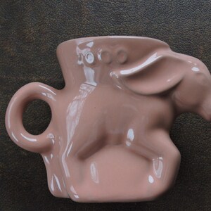 May include: A pink ceramic mug shaped like a donkey with a handle. The mug has three small circles on the top of the head.