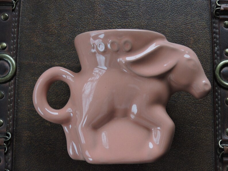 May include: A pink ceramic mug shaped like a donkey with a handle. The mug has three small circles on the top of the head.
