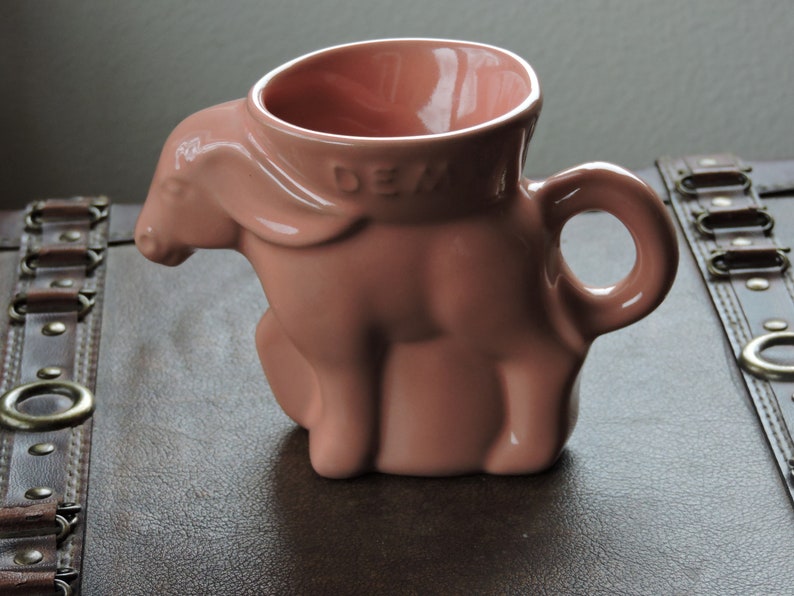 May include: A pink ceramic mug shaped like a pig. The mug has a handle and the word "DEW" is printed on the side.