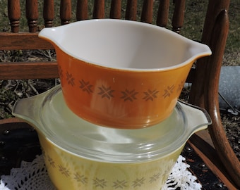 May include: Two vintage Pyrex mixing bowls with a snowflake pattern. The top bowl is orange with brown snowflakes and the bottom bowl is yellow with brown snowflakes.