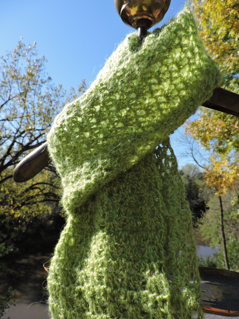 May include: A green crocheted scarf with a delicate openwork pattern and small green beads. The scarf is draped over a metal hook.