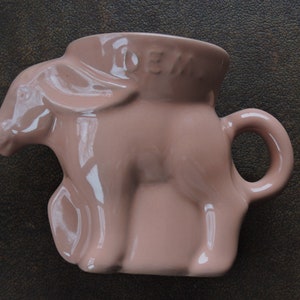 May include: A pink ceramic mug shaped like a donkey with the word "DEN" on the side.
