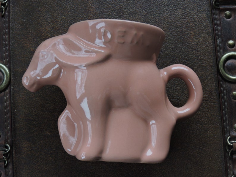 May include: A pink ceramic mug shaped like a donkey with the word "DEN" on the side.