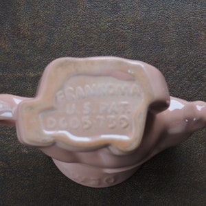 May include: A pink ceramic figurine of a hand holding a pitcher. The bottom of the figurine has the text "FRANKLIN U.S. PAT. D605739".