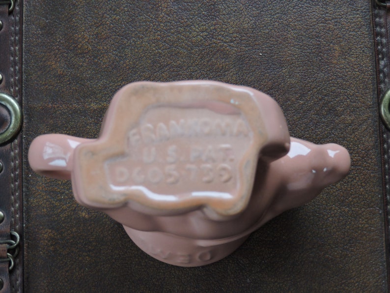 May include: A pink ceramic figurine of a hand holding a pitcher. The bottom of the figurine has the text "FRANKLIN U.S. PAT. D605739".