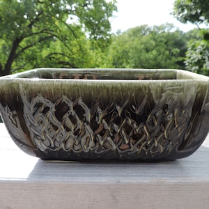 May include: A rectangular ceramic planter with a dark green and brown glaze. The planter has a textured, woven-like pattern on the sides. The background features a blurred view of green trees and a wooden surface.