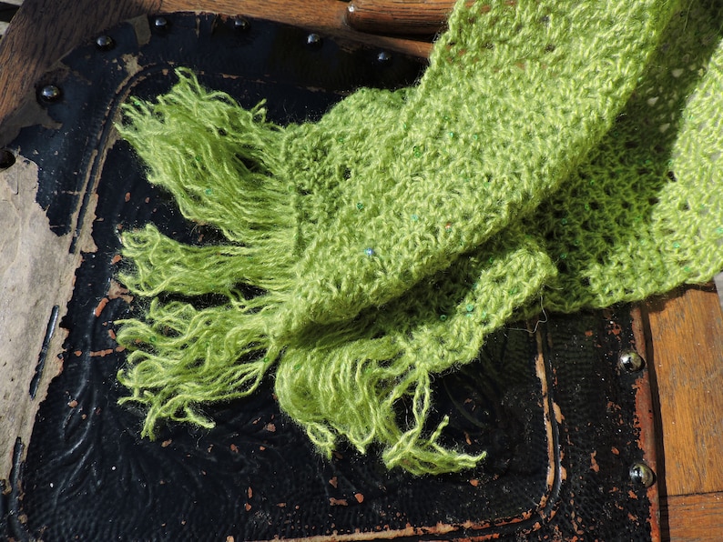May include: A green crocheted scarf with a fringe. The scarf is draped over a wooden chair with a black leather seat.