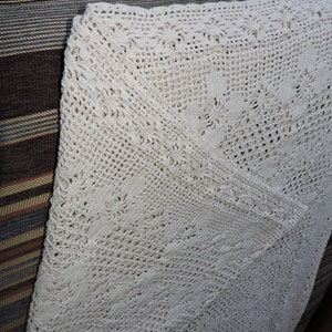May include: Cream-colored, handmade crochet blanket with intricate floral and geometric patterns. The blanket is folded, revealing the detailed openwork design and textured surface. The blanket is a cozy home decor item.