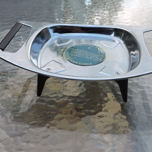 May include: A silver serving tray with black handles and three legs. The tray has a round label in the center that reads "Corning Ware 4 Hour Candle".