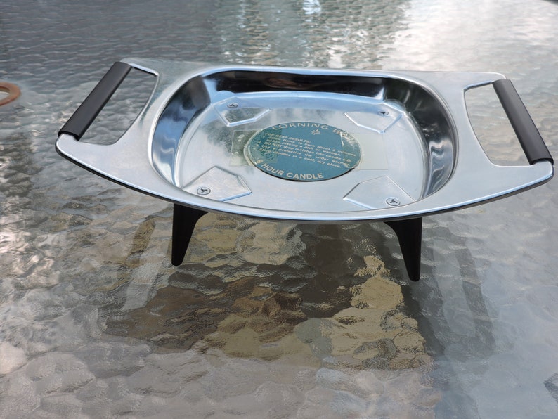 May include: A silver serving tray with black handles and three legs. The tray has a round label in the center that reads "Corning Ware 4 Hour Candle".