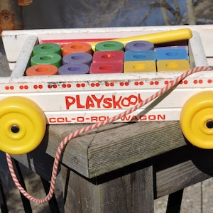 May include: A vintage Playskool Col-O-Rol Wagon toy with a white wooden body, yellow wheels, and a red and white rope. The wagon is filled with colorful wooden blocks.